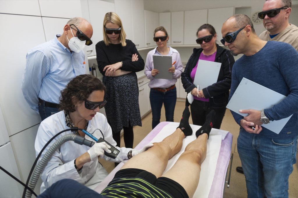 Kursteilnehmerinnen und Kursteilnehmer mit Laserschutzbrillen verfolgen eine Laserbehandlung am Bein